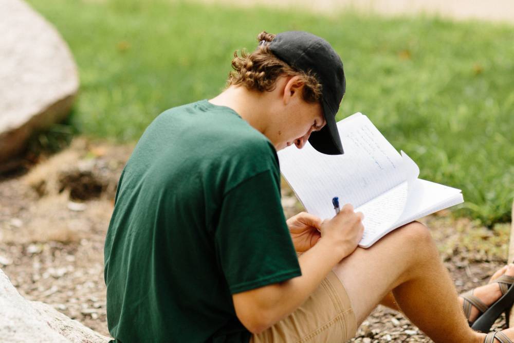 A student sits on the campus lawn writing in a notebook